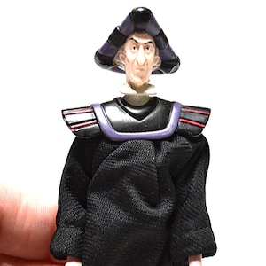 May include: A figurine of a character with a stern expression, wearing a black robe and a tall, triangular hat with purple and black stripes. The figure has a white collar and a black shoulder piece with red accents.