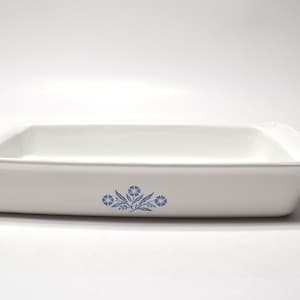May include: A rectangular white ceramic baking dish with a flared rim and a centered blue floral pattern on the front side. The dish features a smooth, glossy finish and a deep interior basin, designed for oven use and food preparation.