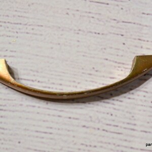 May include: A curved, copper-toned cabinet pull with a slightly arched design. The handle has a smooth, metallic finish and is set against a white, textured background. The pull is designed for furniture and has a vintage aesthetic.