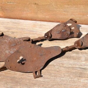 May include: A pair of antique metal ice skates featuring a heavily rusted, weathered brown finish. Each skate consists of a flat, teardrop-shaped footplate with a small central screw mechanism and a hinged, slotted heel plate connected by a metal rod.