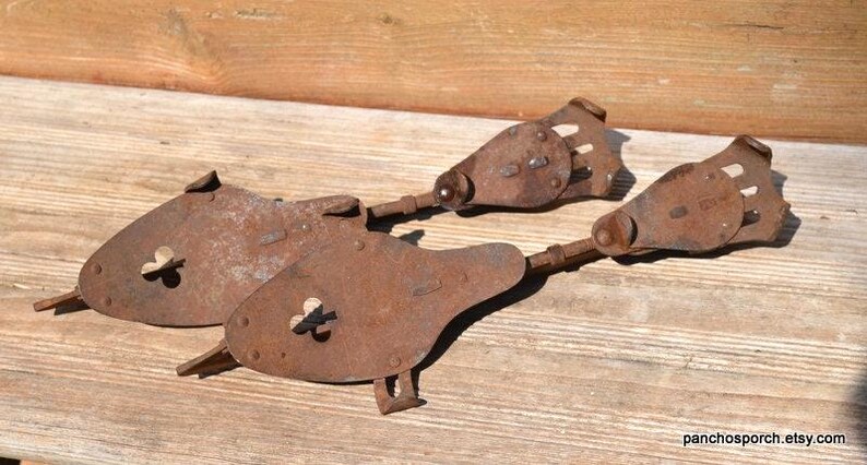 May include: A pair of antique metal ice skates featuring a heavily rusted, weathered brown finish. Each skate consists of a flat, teardrop-shaped footplate with a small central screw mechanism and a hinged, slotted heel plate connected by a metal rod.