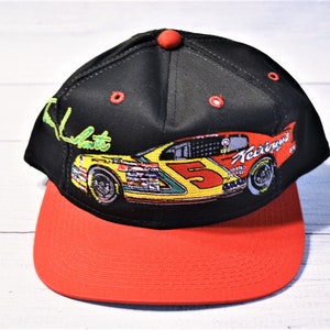May include: Black baseball cap with a red bill and button. The cap features an embroidered race car design with the number 5 and the name "Kellogg's". The cap also has the embroidered signature "Ken Schrader" in green.