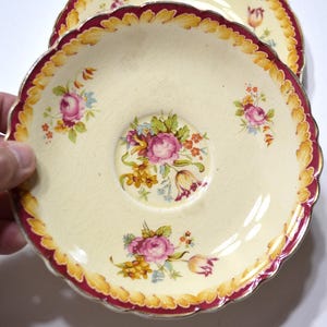 May include: A cream-colored ceramic saucer with a scalloped edge featuring a deep red and yellow border. The center and rim are decorated with floral bouquets of pink roses, yellow flowers, and green leaves, finished with a visible crackle glaze.