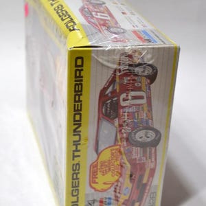Puede incluir: Un modelo en caja de un coche de carreras Folgers Thunderbird. La caja es amarilla y presenta im&aacute;genes del coche en rojo, blanco y azul. La caja est&aacute; envuelta en pl&aacute;stico transparente.