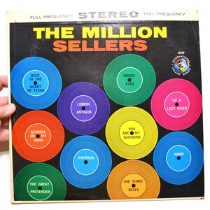 May include: Vintage vinyl record album titled "The Million Sellers." The album cover is black with colorful circular designs, each featuring a song title like "Green Eyes" and "Georgia on My Mind." The album is held by a hand.