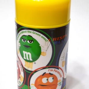 May include: A cylindrical container with a bright yellow lid and a black body. The body features cartoon characters and text, including "What is it about the Green ones?" and "I'm Paranoid!" The container is likely for candy.