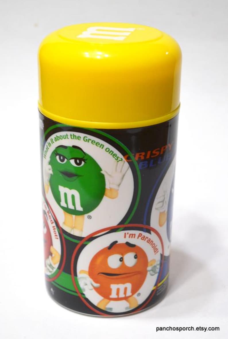 May include: A cylindrical container with a bright yellow lid and a black body. The body features cartoon characters and text, including "What is it about the Green ones?" and "I'm Paranoid!" The container is likely for candy.