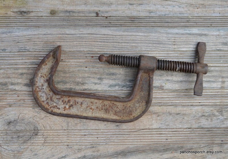 May include: A rusty metal C-clamp with a threaded screw and a wing nut. The clamp is on a wooden surface.