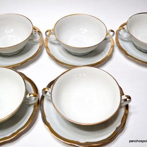 May include: A set of six white porcelain soup bowls with matching saucers. Each bowl has two handles and a gold rim, as does each saucer. The saucers have a scalloped edge. The bowls and saucers are arranged on a white surface.
