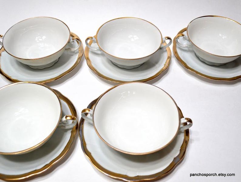 May include: A set of six white porcelain soup bowls with matching saucers. Each bowl has two handles and a gold rim, as does each saucer. The saucers have a scalloped edge. The bowls and saucers are arranged on a white surface.