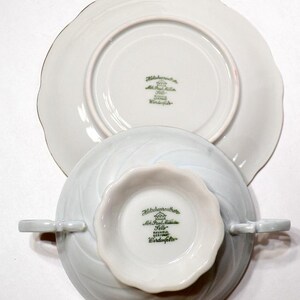 May include: A white porcelain teacup and saucer set with a leaf-like design. The teacup has two small handles and a scalloped rim. The saucer is also white with a scalloped edge. The bottom of both pieces have green text that reads "Hutschenreuther".