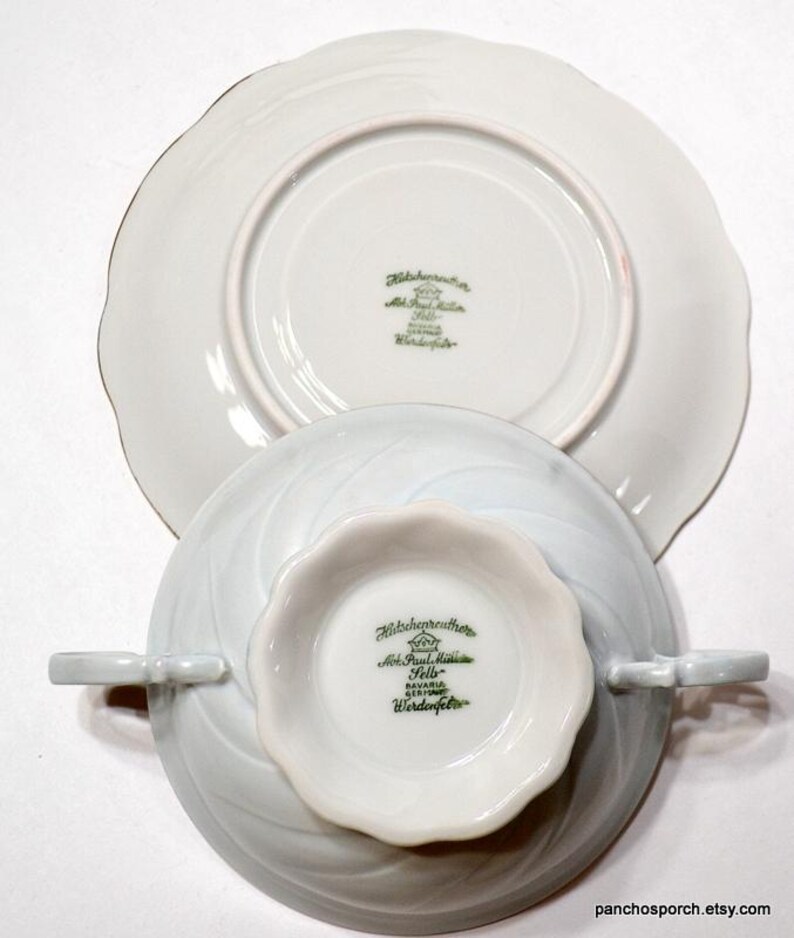 May include: A white porcelain teacup and saucer set with a leaf-like design. The teacup has two small handles and a scalloped rim. The saucer is also white with a scalloped edge. The bottom of both pieces have green text that reads "Hutschenreuther".