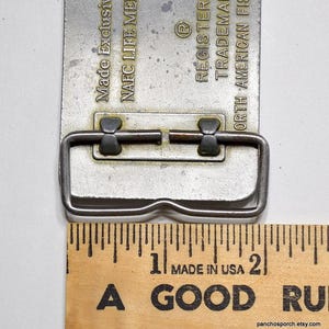 May include: A silver belt buckle with a metal loop and two small metal tabs. The buckle is engraved with the text "Made Exclusive", "NAFC LIFE MEN", "REGISTERED", "TRADEMARK", and "NORTH AMERICAN FISHING". The buckle is measured against a wooden ruler.