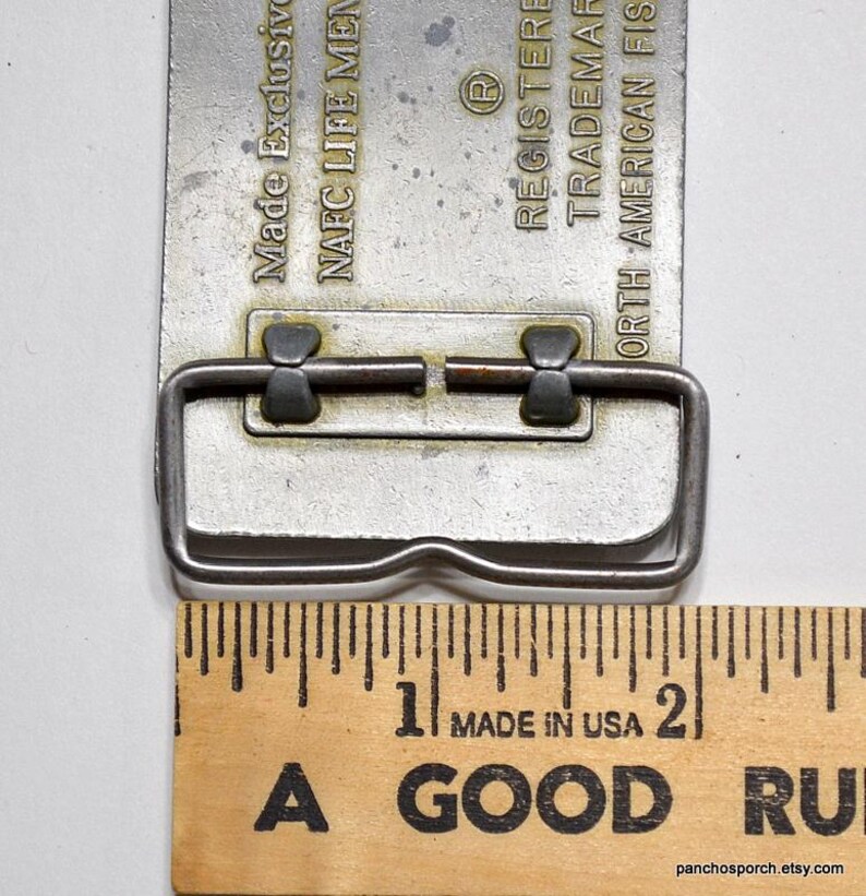 May include: A silver belt buckle with a metal loop and two small metal tabs. The buckle is engraved with the text "Made Exclusive", "NAFC LIFE MEN", "REGISTERED", "TRADEMARK", and "NORTH AMERICAN FISHING". The buckle is measured against a wooden ruler.
