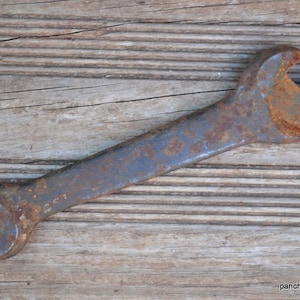 May include: A vintage, rusty wrench with a curved handle and open ends. The metal tool shows signs of age and wear, with a weathered, textured surface and areas of rust. The wrench is set against a backdrop of weathered wooden planks.