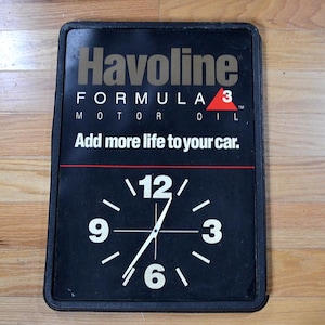 May include: Black rectangular clock with the Havoline Formula 3 Motor Oil logo. The clock face displays the numbers 12, 3, 6, and 9 in white, with white hands. The text "Add more life to your car" is also visible.