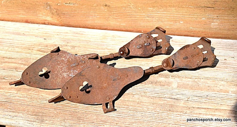 May include: Two antique metal ice skates with a heavy rusted finish. Each skate features a teardrop-shaped plate with a clover-shaped cutout, a long metal runner, and a mechanical heel adjustment mechanism with a series of small rectangular slots.