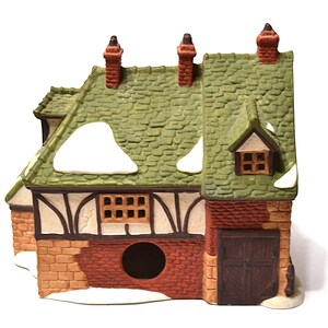 May include: A ceramic miniature house with a green shingled roof, brick chimney, and snow accents. The house features a brown door, windows, and a brick and stucco facade. A charming decorative piece.