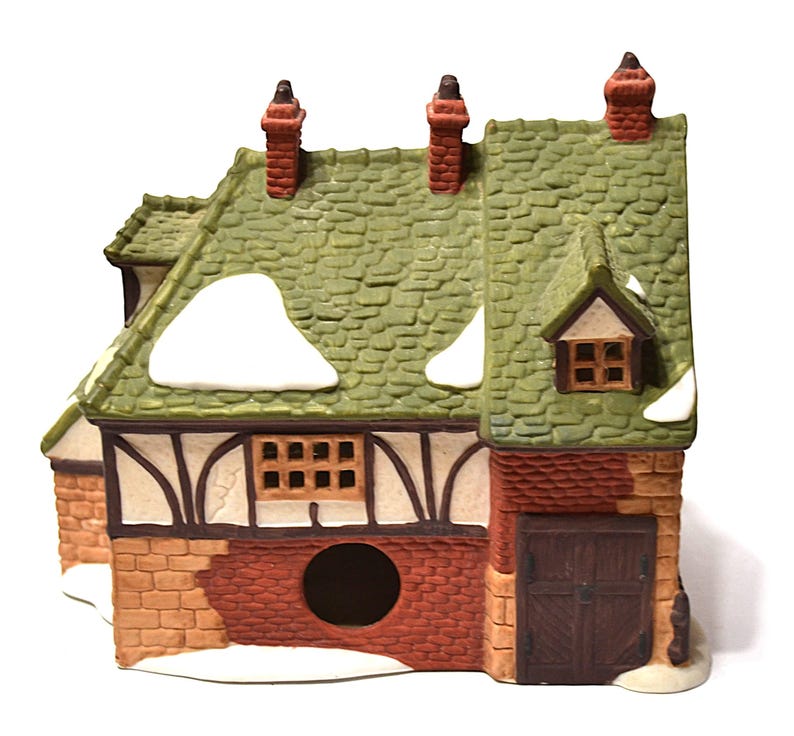 May include: A ceramic miniature house with a green shingled roof, brick chimney, and snow accents. The house features a brown door, windows, and a brick and stucco facade. A charming decorative piece.