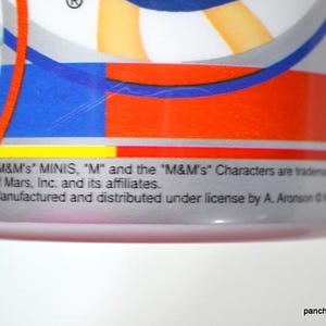 May include: Close-up of a cylindrical container with a colorful design featuring red, blue, and orange stripes. The text "M&M's MINIS" is visible, along with other text indicating trademarks and licensing information. The bottom of the container is pink.