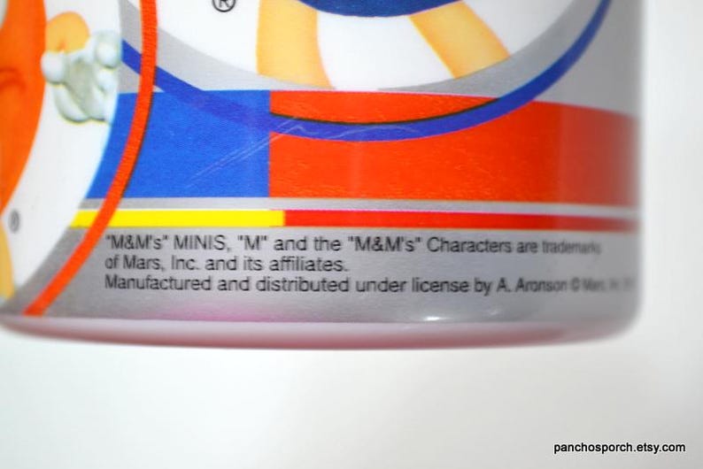 May include: Close-up of a cylindrical container with a colorful design featuring red, blue, and orange stripes. The text "M&M's MINIS" is visible, along with other text indicating trademarks and licensing information. The bottom of the container is pink.