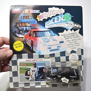 May include: A vintage NASCAR Roaring Racer toy car set. The packaging features illustrations of race cars with numbers 44, 76, and 51. Includes a die-cast stock car, collector's card, and real engine sounds. The set includes a Dale Earnhardt card.
