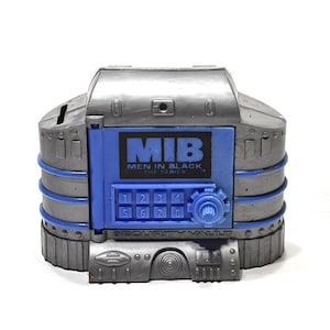 May include: A silver and blue plastic Men in Black The Series security vault coin bank. The front features a blue keypad with numbers one through eight, a circular dial, and the text MIB MEN IN BLACK THE SERIES printed above the keypad.