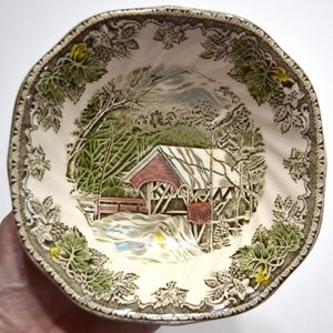 May include: A ceramic bowl with a scalloped edge featuring a transferware print of a red covered bridge in a snowy landscape. The border is decorated with green leaves and white flowers on a cream background, creating a detailed scene.