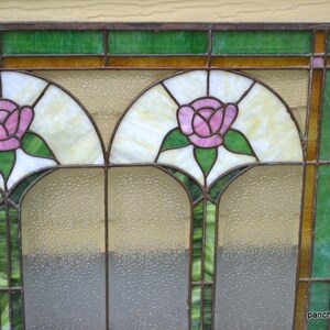 Op de afbeelding: Een glas-in-loodraam met een geometrisch ontwerp met twee roze rozen met groene bladeren. Het raam heeft een bruin houten frame en is geplaatst tegen een lichtbruine muur.