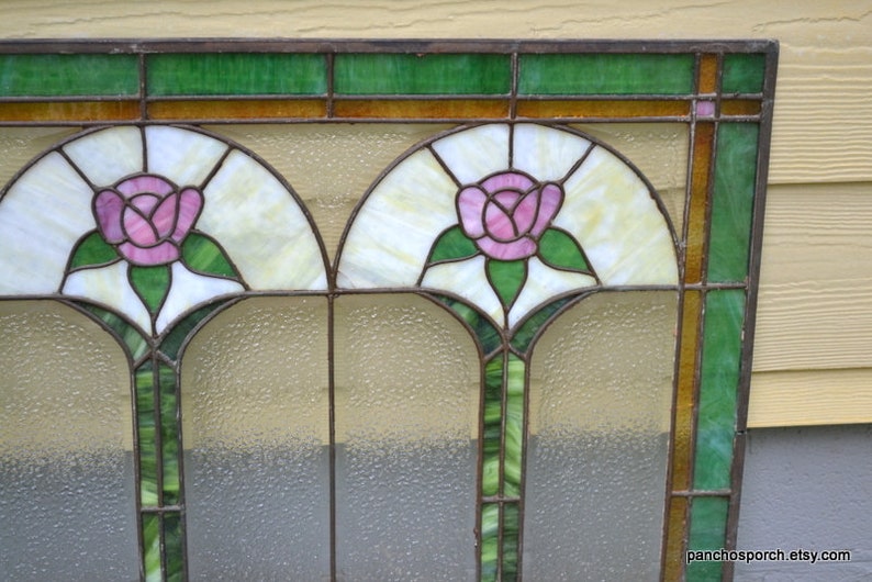 Op de afbeelding: Een glas-in-loodraam met een geometrisch ontwerp met twee roze rozen met groene bladeren. Het raam heeft een bruin houten frame en is geplaatst tegen een lichtbruine muur.