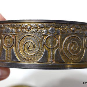 Op de afbeelding: Decoratieve metalen band met een ingewikkeld ontwerp met goud- en zilverkleuren. De band heeft een herhalend patroon van spiralen en gestileerde bloemelementen. Het item wordt door een hand vastgehouden.