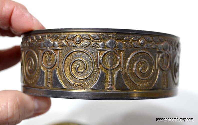 Op de afbeelding: Decoratieve metalen band met een ingewikkeld ontwerp met goud- en zilverkleuren. De band heeft een herhalend patroon van spiralen en gestileerde bloemelementen. Het item wordt door een hand vastgehouden.