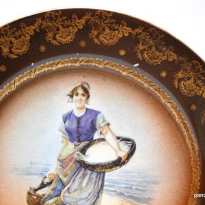 Op de afbeelding: Antieke decoratieve schotel met een bruine rand met gouden bloemmotieven en een centraal beeld van een vrouw die manden draagt ​​bij de zee. De vrouw draagt ​​een blauwe jurk en een witte hoofdbedekking.
