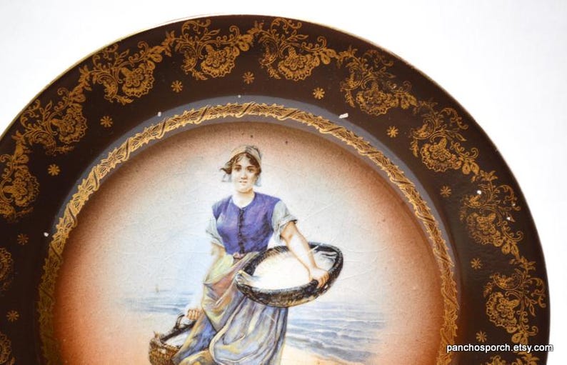 Op de afbeelding: Antieke decoratieve schotel met een bruine rand met gouden bloemmotieven en een centraal beeld van een vrouw die manden draagt ​​bij de zee. De vrouw draagt ​​een blauwe jurk en een witte hoofdbedekking.