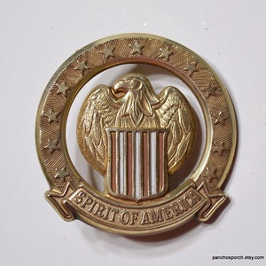 May include: A circular gold-toned metal pin featuring an eagle with wings spread above a shield with red, white, and blue vertical stripes. The eagle is surrounded by a ring of stars, and a banner at the bottom reads SPIRIT OF AMERICA.