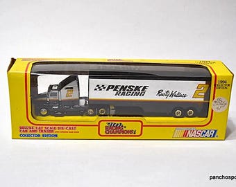 Op de afbeelding: Een model van een Penske Racing-vrachtwagen met nummer 2 en de naam Rusty Wallace. Het model zit in een gele doos met de tekst "NASCAR" en "Collector Edition". De vrachtwagen is zwart, wit en goud.