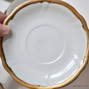 May include: White saucer with a gold rim and a scalloped edge. The saucer has a simple, elegant design with a slightly raised center. The gold rim adds a touch of luxury to the classic white porcelain.
