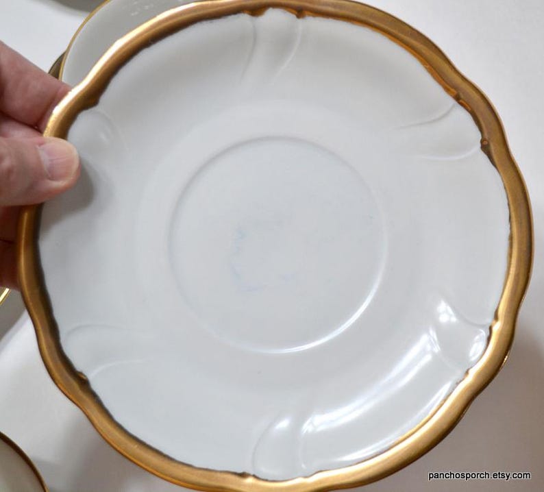 May include: White saucer with a gold rim and a scalloped edge. The saucer has a simple, elegant design with a slightly raised center. The gold rim adds a touch of luxury to the classic white porcelain.