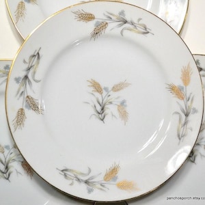 May include: White ceramic plates with a gold rim and wheat stalk design. The wheat stalks are in shades of gold and brown, with accents of blue and gray. The plates are arranged in a stack.