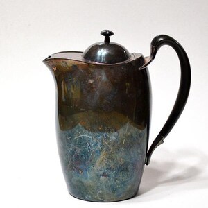 May include: Vintage silver-plated pitcher with a black handle and lid. The pitcher has a classic design with a spout for pouring. The metal has a patina with shades of blue and green. A decorative piece for serving beverages.