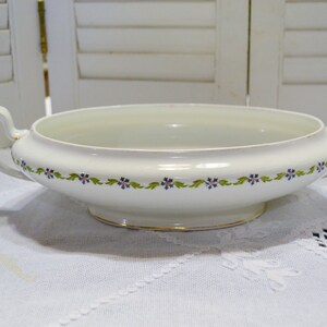 May include: A white ceramic serving bowl with two handles. The bowl has a floral design of blue flowers and green leaves around the rim. The base has a gold trim. The bowl is on a white tablecloth.