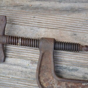 May include: A rusty metal C-clamp with a threaded screw and a handle. The clamp is on a wooden surface.