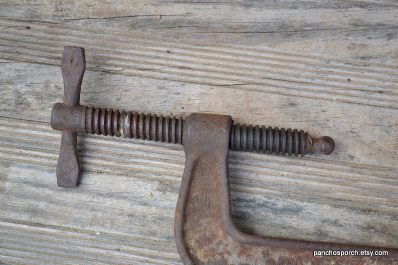May include: A rusty metal C-clamp with a threaded screw and a handle. The clamp is on a wooden surface.