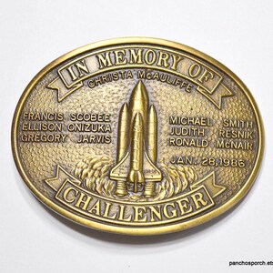 May include: A gold-toned metal belt buckle commemorating the Space Shuttle Challenger disaster. The buckle features an image of the Challenger space shuttle, the names of the crew members, and the date of the disaster, January 28, 1986.