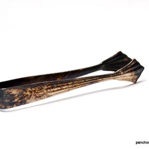 May include: Antique silver-toned sugar tongs with a decorative floral pattern. The tongs have a classic design with a curved handle and a pinching mechanism. The metal has a tarnished appearance, adding to its vintage charm.