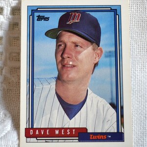 May include: A Topps baseball card featuring Dave West of the Minnesota Twins. The card has a blue border, with the Topps logo at the top. West is wearing a Twins cap and pinstripe jersey. The card's text includes "DAVE WEST" and "Twins".