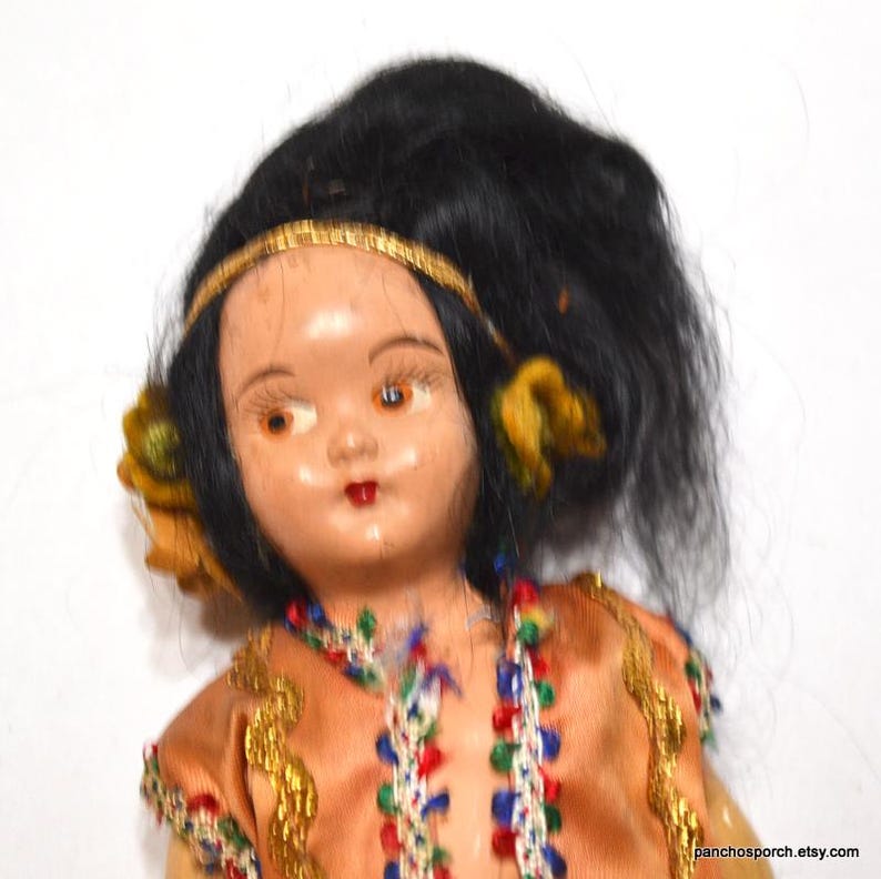 Puede incluir: Mu&ntilde;eca vintage con cabello negro, ojos marrones y un vestido color melocot&oacute;n. La mu&ntilde;eca tiene una diadema dorada y detalles amarillos en el cabello. El vestido est&aacute; adornado con ribetes dorados y cuentas de colores. La cara de la mu&ntilde;eca tiene una expresi&oacute;n pintada.