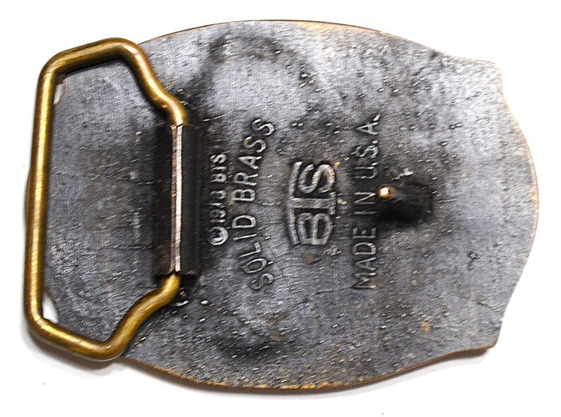 May include: Vintage solid brass belt buckle with a rectangular frame and an oval body. The buckle is marked "BIS SOLID BRASS" and "MADE IN U.S.A.". The metal has a tarnished, aged appearance.