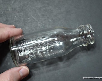 May include: A clear glass bottle with the embossed text "A. Florence & Co." is held in a hand. The bottle has a rounded body and a narrow neck. The bottle is empty and appears to be vintage.