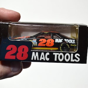 May include: A black and orange die-cast stock car toy in its original packaging. The car features the number 28 and Mac Tools branding. The black box displays the number 28 in large red font and the text Mac Tools in white, with a clear plastic window.